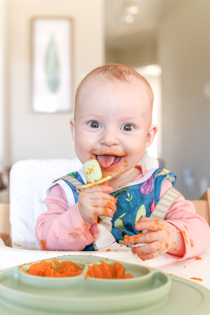 a 6 month old baby wearing a blue bib eating solid food with a spoon