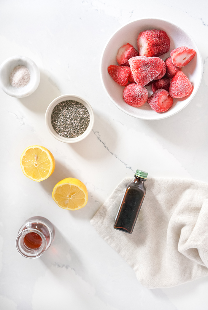 Ingredients for strawberry chia jam laid out on a white table, including frozen strawberries, chia seeds, lemon, maple syrup and vanilla essence