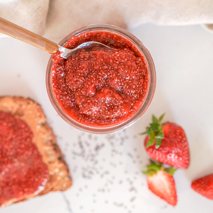 A pot of strawberry chia jam sitting on a white table next to strawberries, chia seeds and a piece of toast spread with chia jam