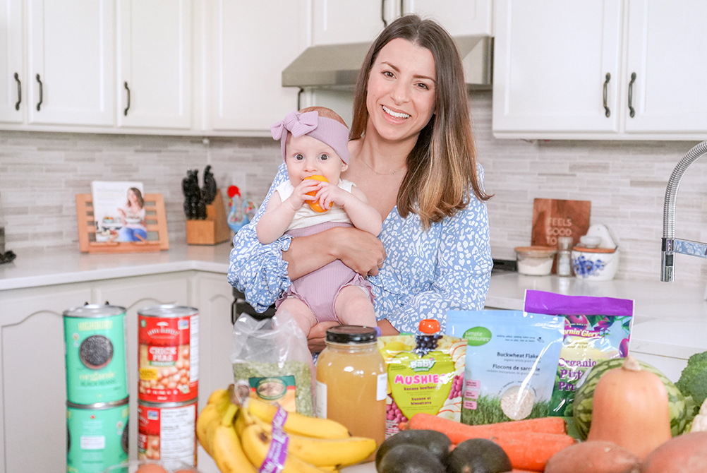Brunette woman holding her 7 month old baby and showing off baby led weaning foods in her kitchen