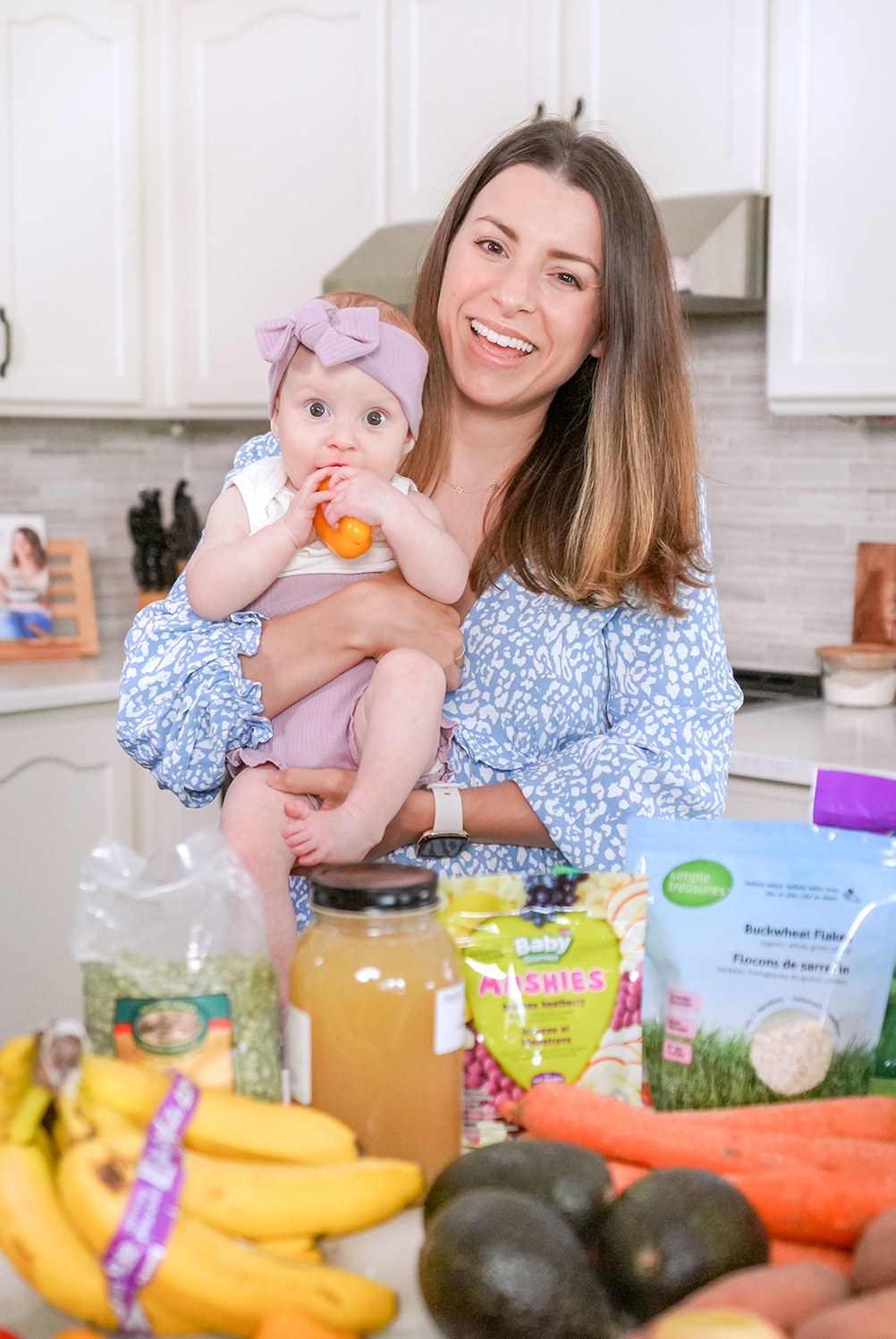 Brunette woman holding her 7 month old baby and showing off baby led weaning foods in her kitchen