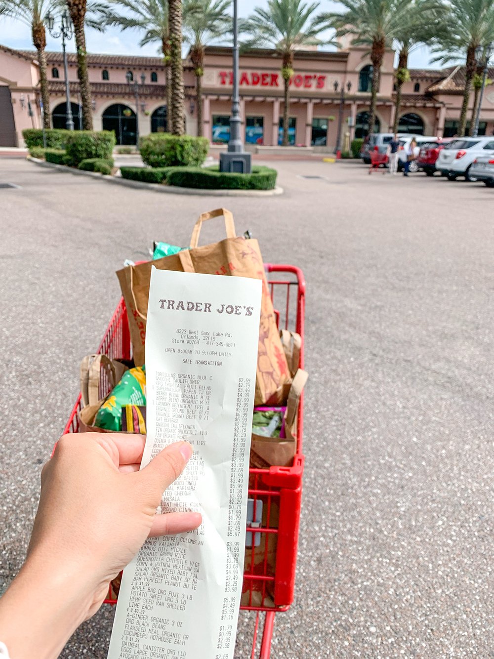 a hand holding up a Trader Joe's receipt in front of a shopping cart full of food