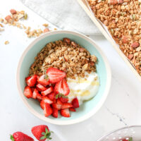 a bowl of yogurt parfait and granola on a white table, with granola and strawberries next to it