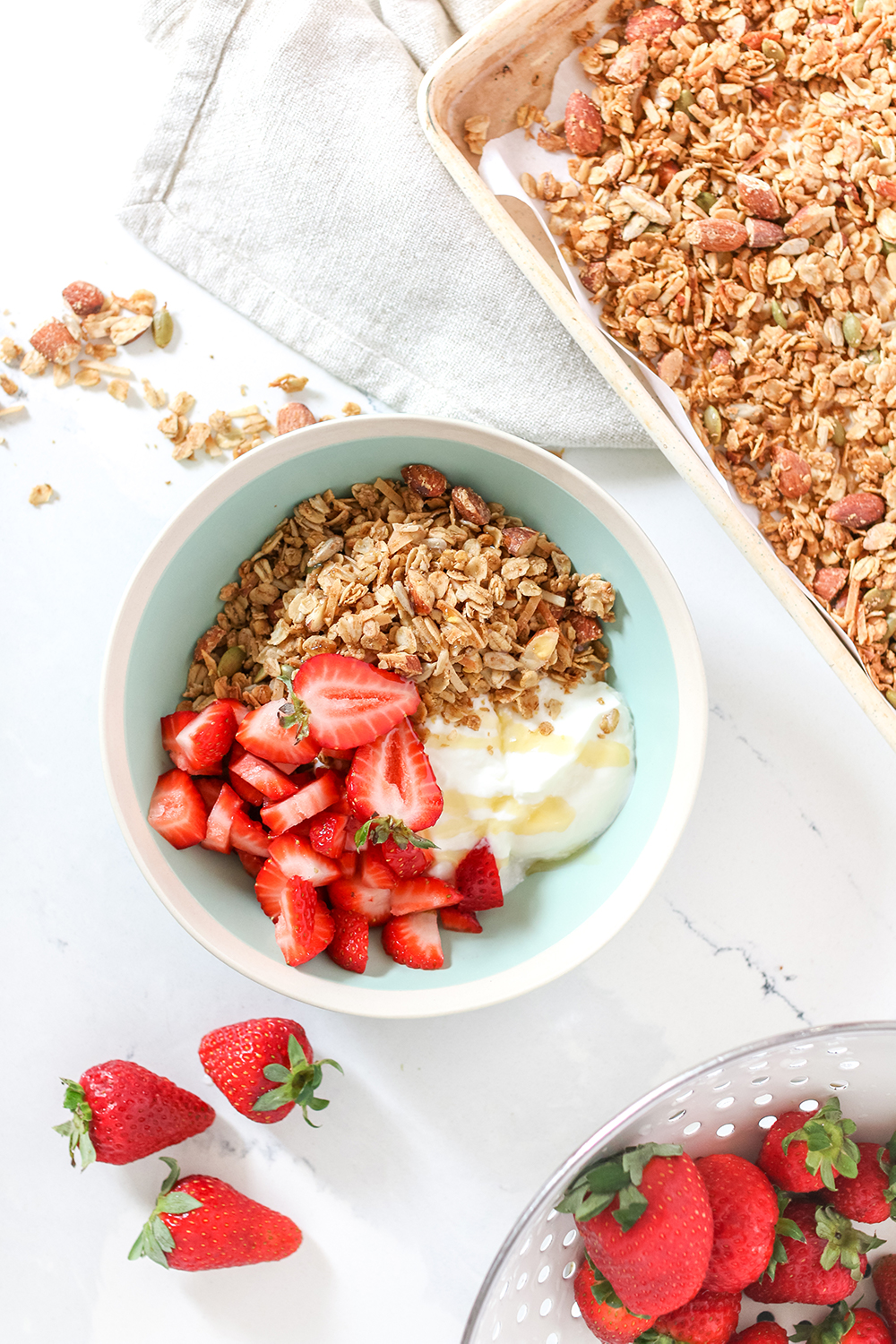 a bowl of yogurt parfait and granola on a white table, with granola and strawberries next to it