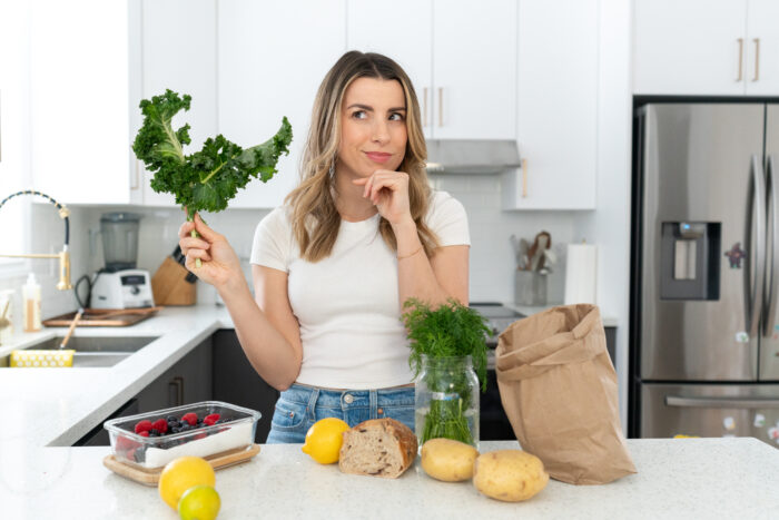 A lady with brown hair standing behind her white kitchen counter with a lot of fruit and vegetables on the counter.